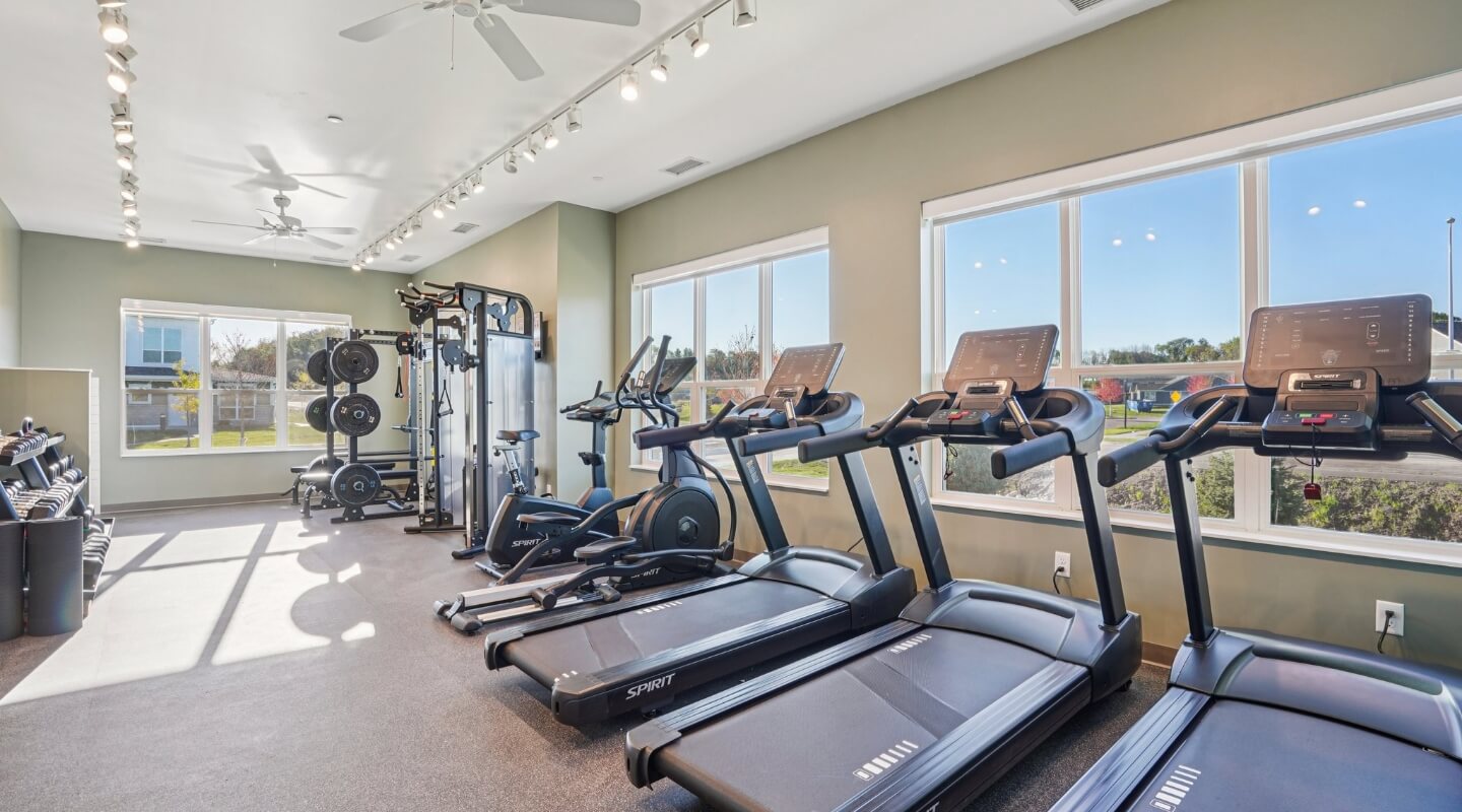 Bright fitness room with treadmills, weight machines, and large windows overlooking the community.
