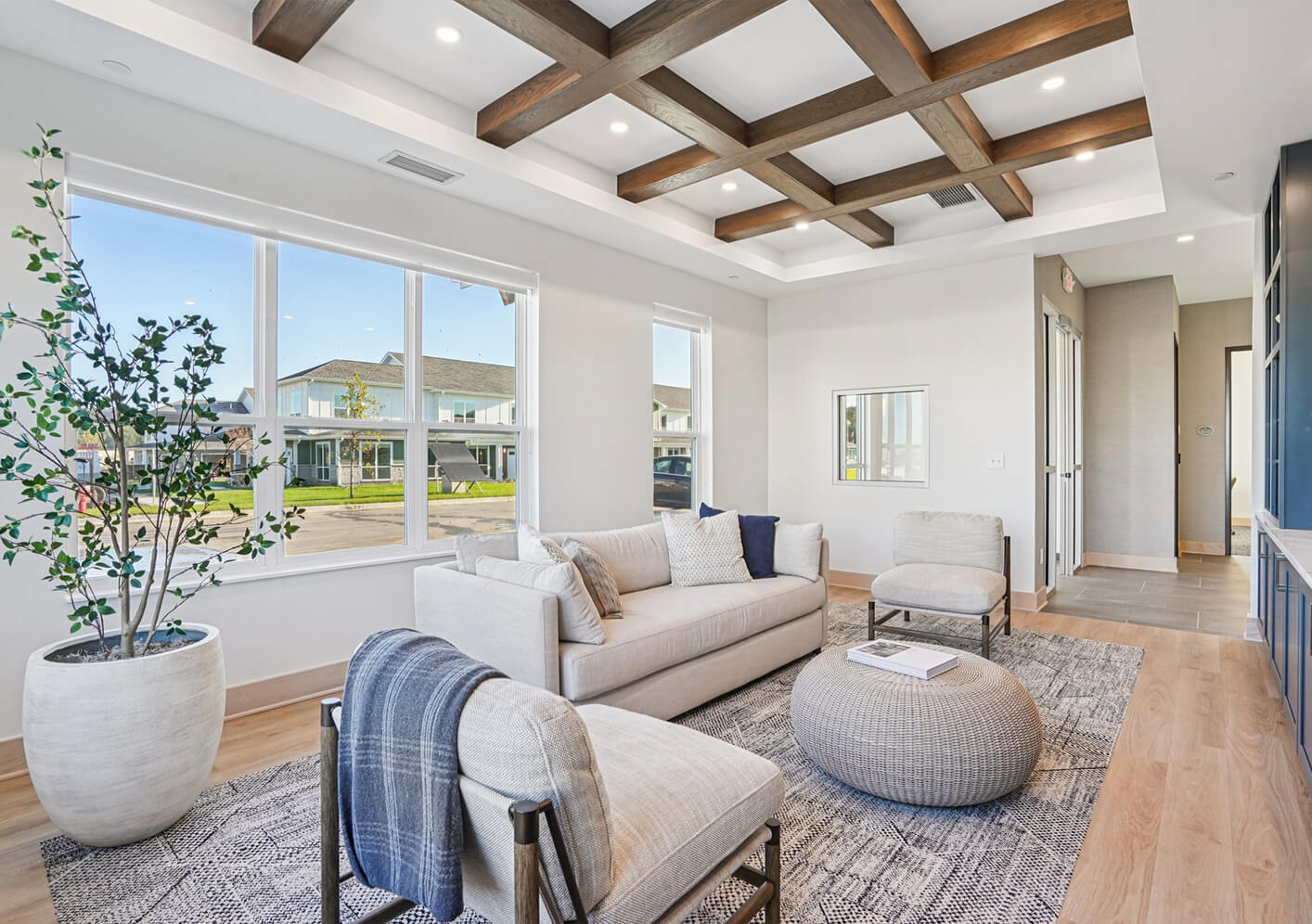 Bright modern living room with large windows, neutral furniture, coffered wood ceiling, and natural light streaming in.