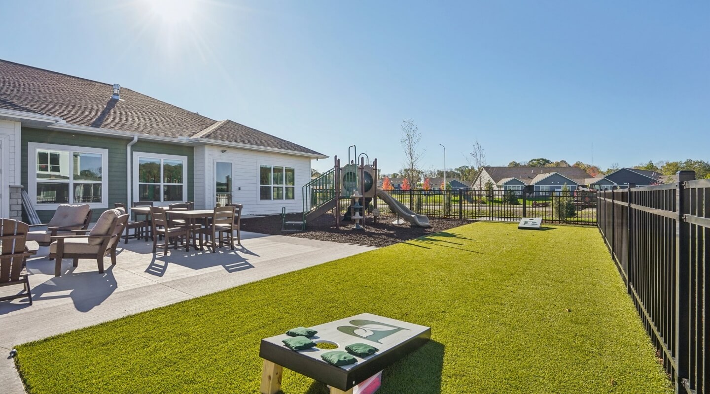 Outdoor patio area with seating, cornhole boards, and playground under a sunny sky.