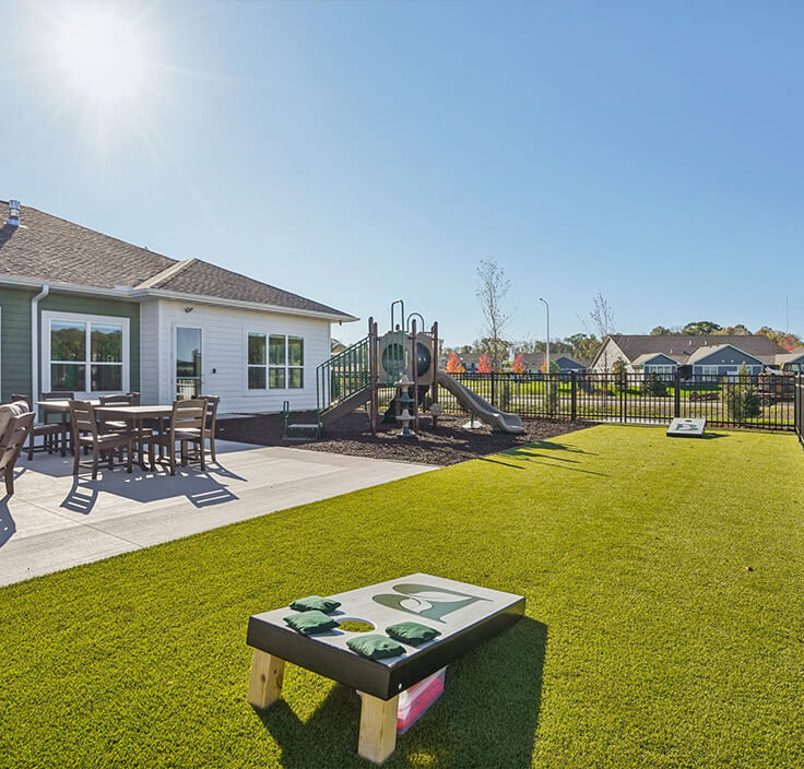 Outdoor community area with playground, patio seating, and cornhole game under a bright sunny sky.